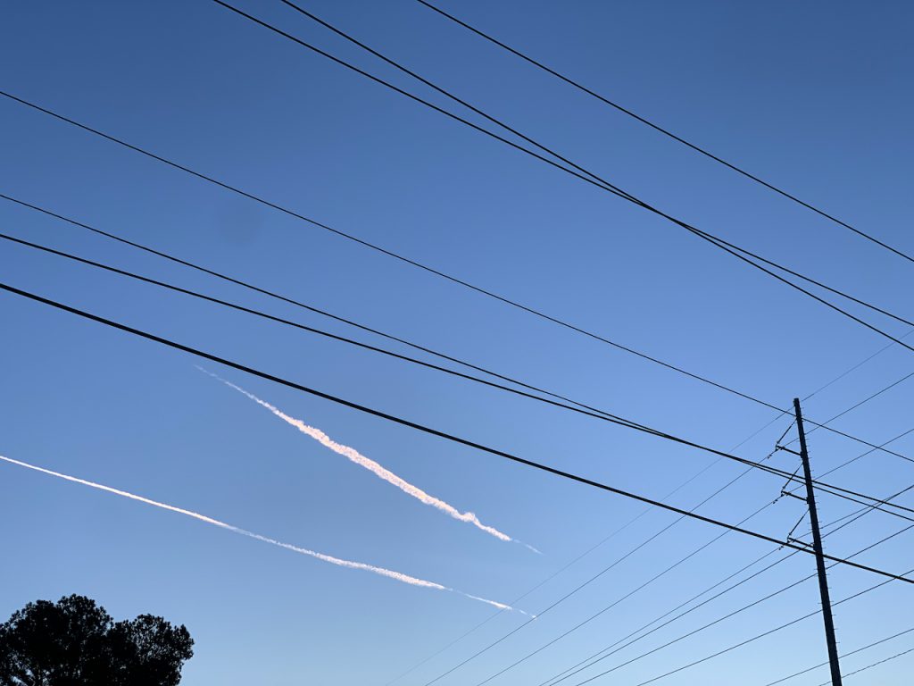 The sky above is depicted. The top of a tree is in the lower left corner. Going across the screen are two contrails from jets overhead, they are already dissolving. Utility wires go left to right across the screen, also in the right corner is another cross section of many utility wires. The sky is blue with a hint of purple.